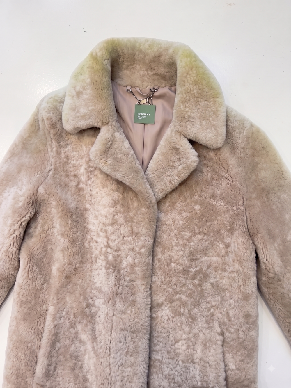 A light beige faux fur coat with a wide collar, laid flat against a plain white background. The interior lining is a dusty pink color, and a green 'L'Envers' brand tag is visible at the collar.