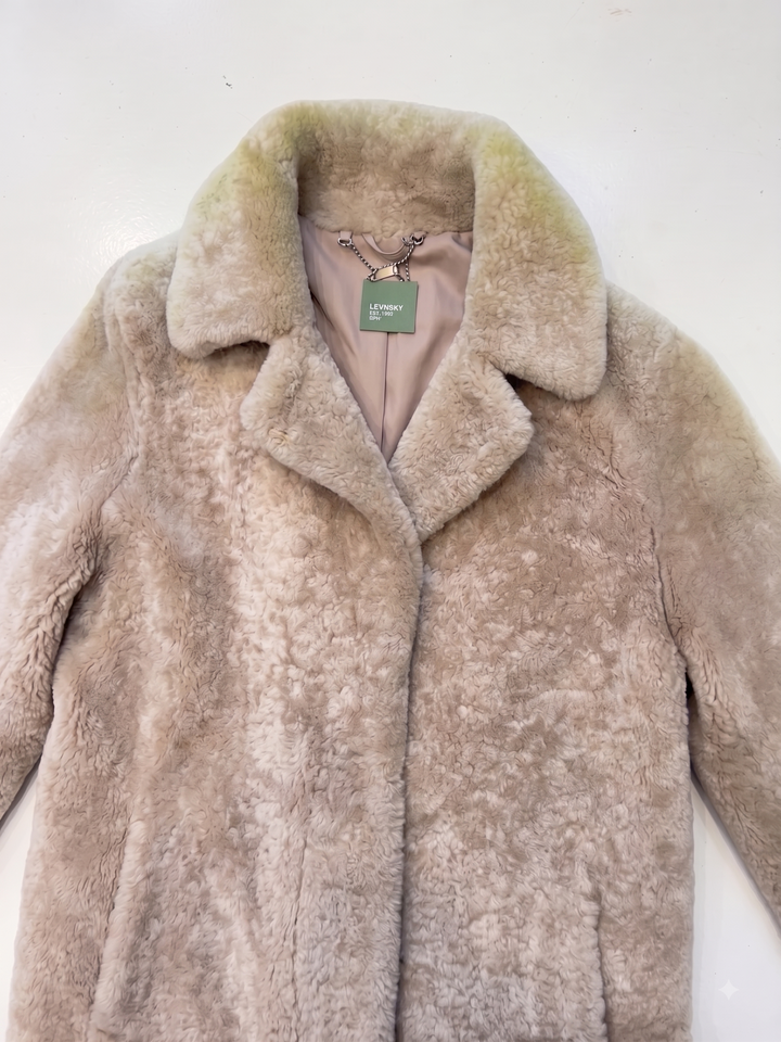 A light beige faux fur coat with a wide collar, laid flat against a plain white background. The interior lining is a dusty pink color, and a green 'L'Envers' brand tag is visible at the collar.