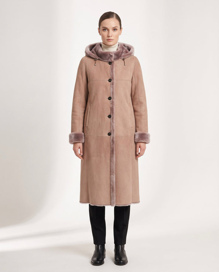 A woman stands facing forward wearing a long taupe-brown shearling-style coat with a fuzzy taupe hood and matching cuff trim, black pants, and black boots against a plain light studio background.
