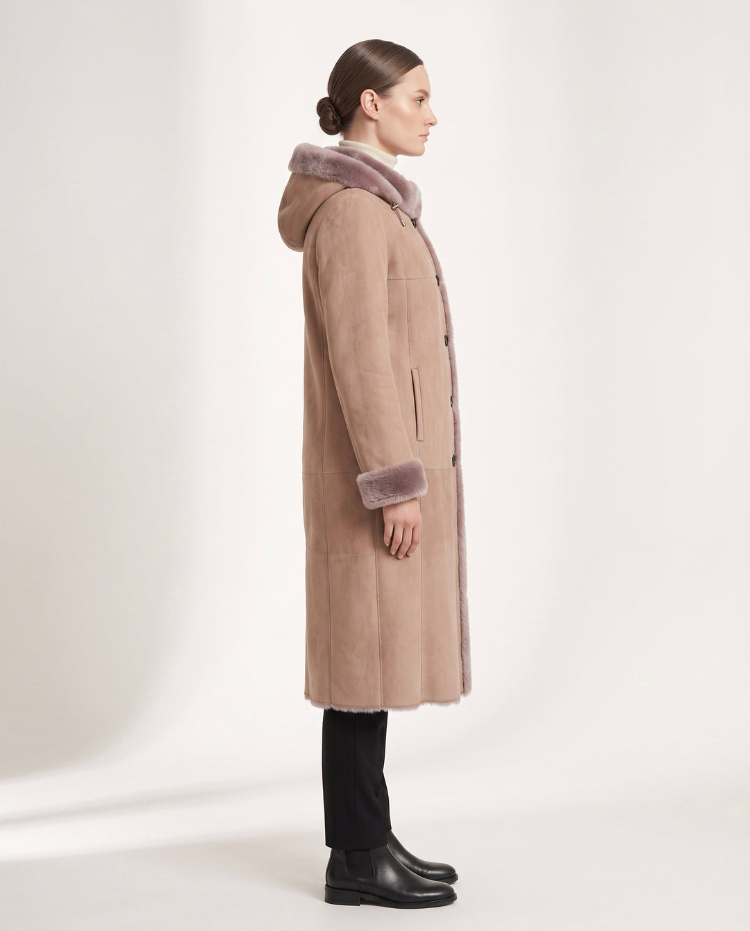 A woman in profile wearing a mid-length beige shearling-style coat with a fuzzy lavender-gray hood and cuffs, paired with black pants and black boots, against a clean off-white studio background.