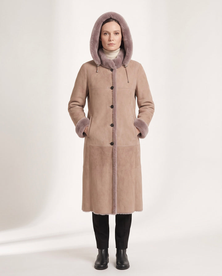 A woman stands against a plain light studio background wearing a long taupe-brown shearling-style coat with a soft mauve-pink hood and trim, black leggings, and black boots.