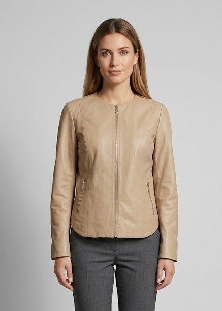 A woman stands against a light gray studio background wearing a tan leather-look zip-front jacket, dark gray tailored trousers, and black heels.