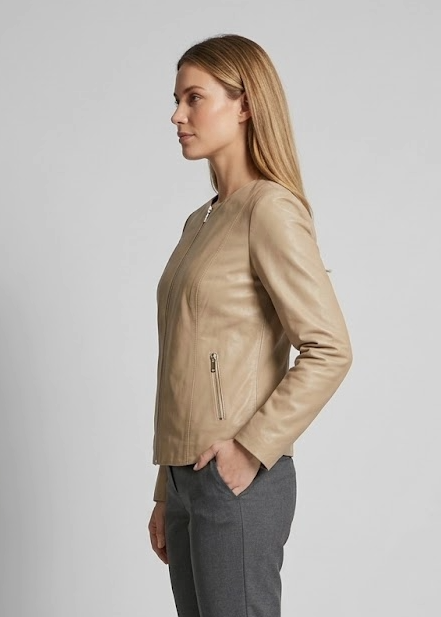 A woman in side profile wearing a beige leather jacket, dark gray trousers, and black heels, standing against a light gray studio background.